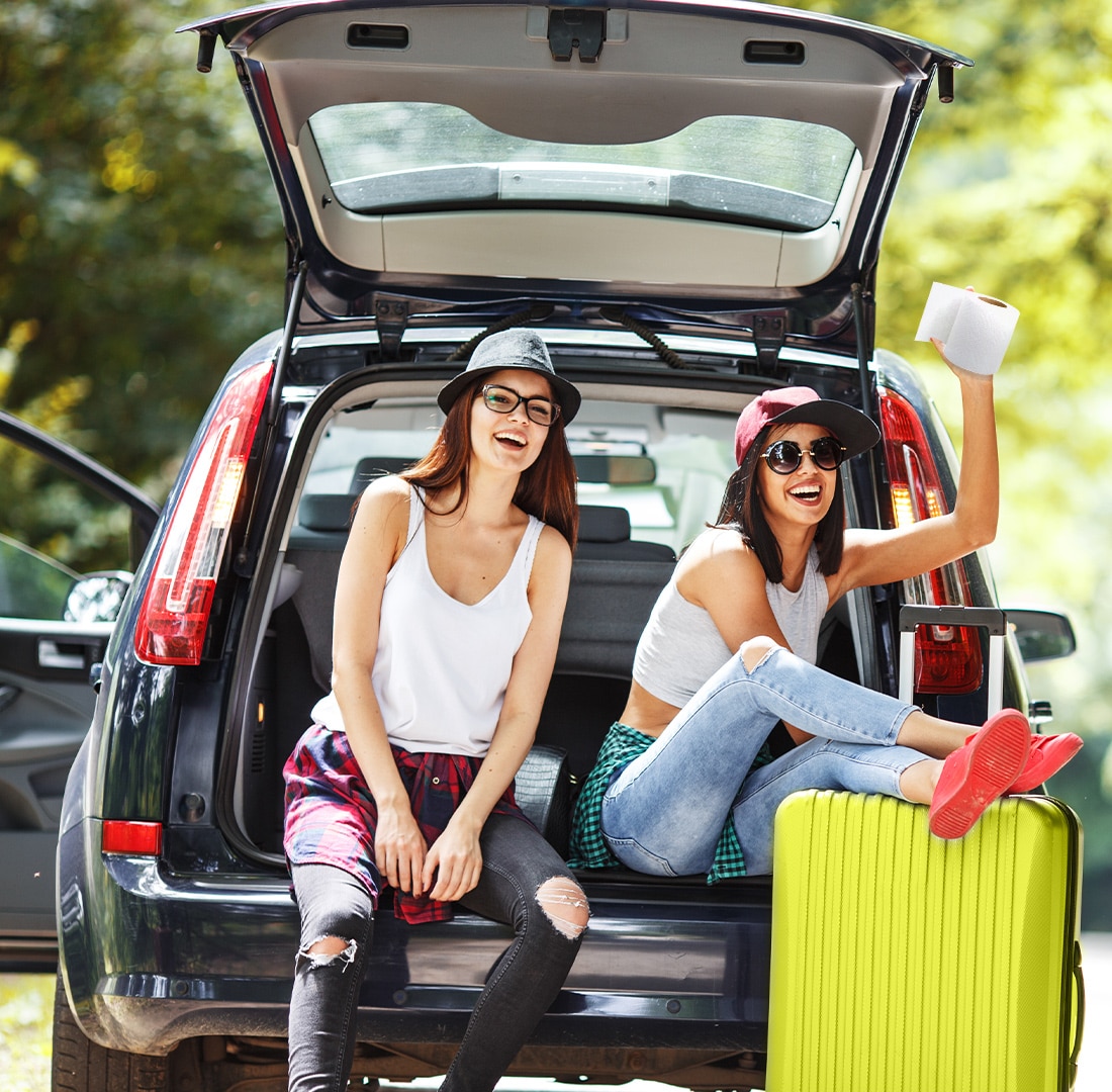 Two Gorls sitting on the rear bumper of a car ready for a road trip holding a roll of toilet paper